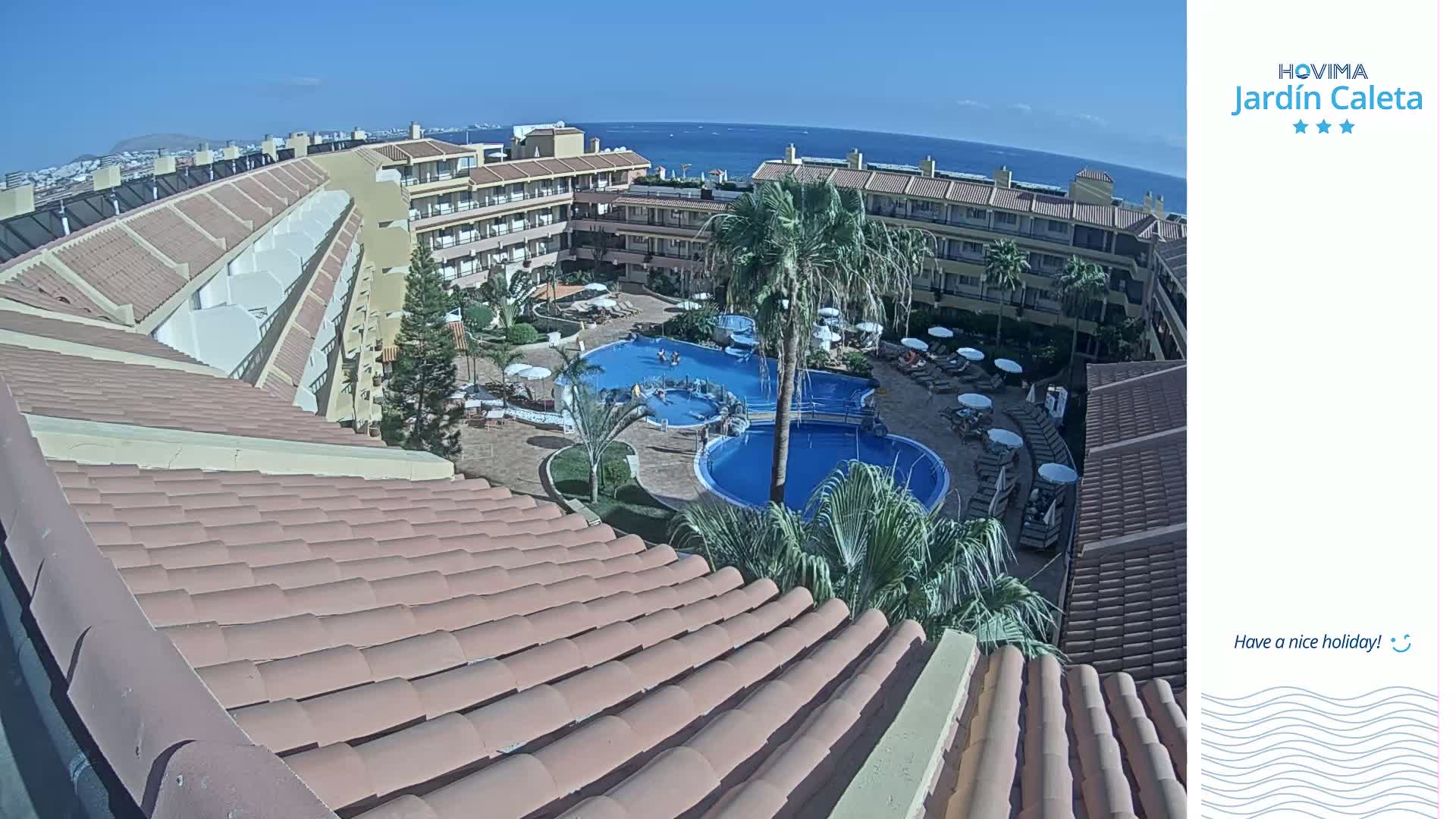 A rooftop view of a resort complex on a sunny day shows multiple swimming pools surrounded by palm trees and lounge chairs, with an ocean visible in the background.