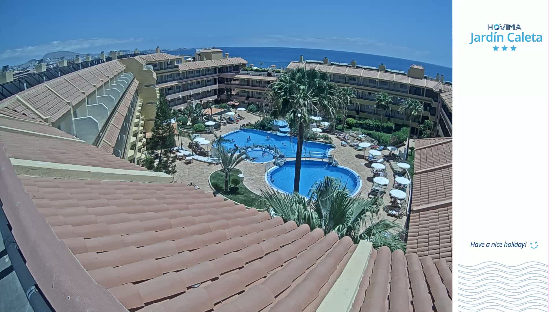 A tiled roof overlooks a sunny resort courtyard with two swimming pools, palm trees, and lounge chairs near the ocean.