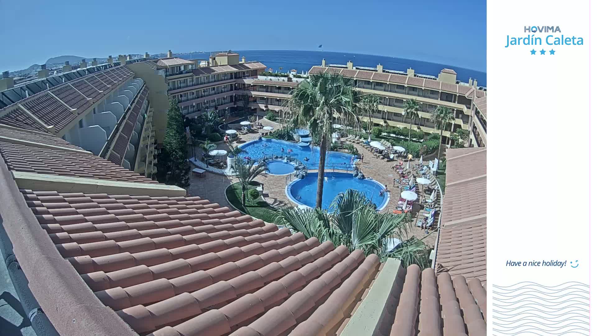 An aerial view of a resort complex on a sunny day shows multiple buildings surrounding a pool area with several people enjoying the water and lounge chairs.