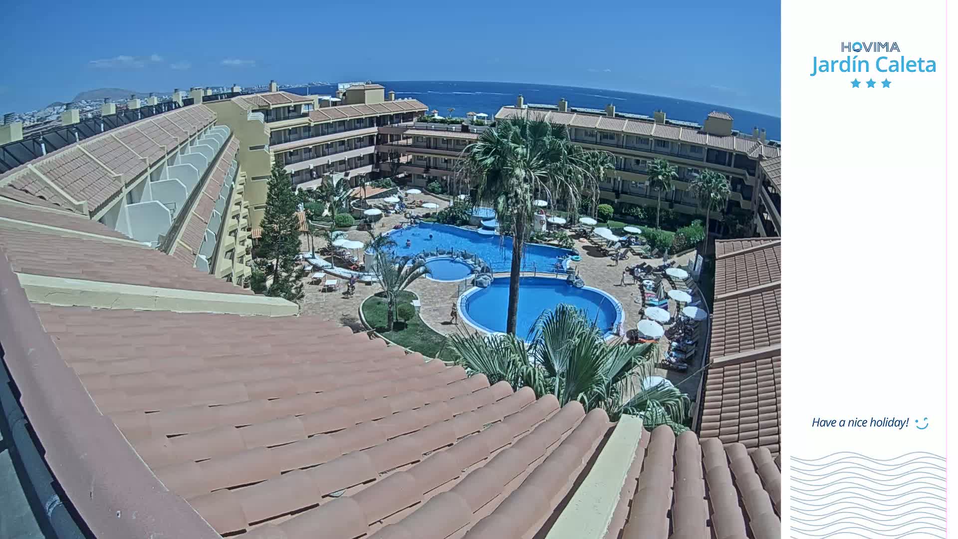 A resort's tiled roof overlooks a courtyard with two swimming pools, surrounded by palm trees, lounge chairs, and people on a sunny day.