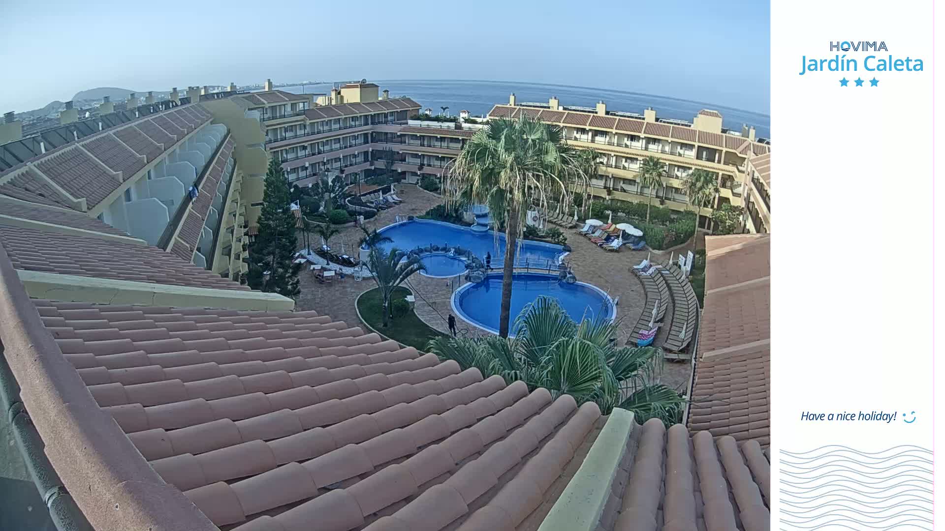 A resort with a large pool is viewed from a rooftop, under a clear sky.