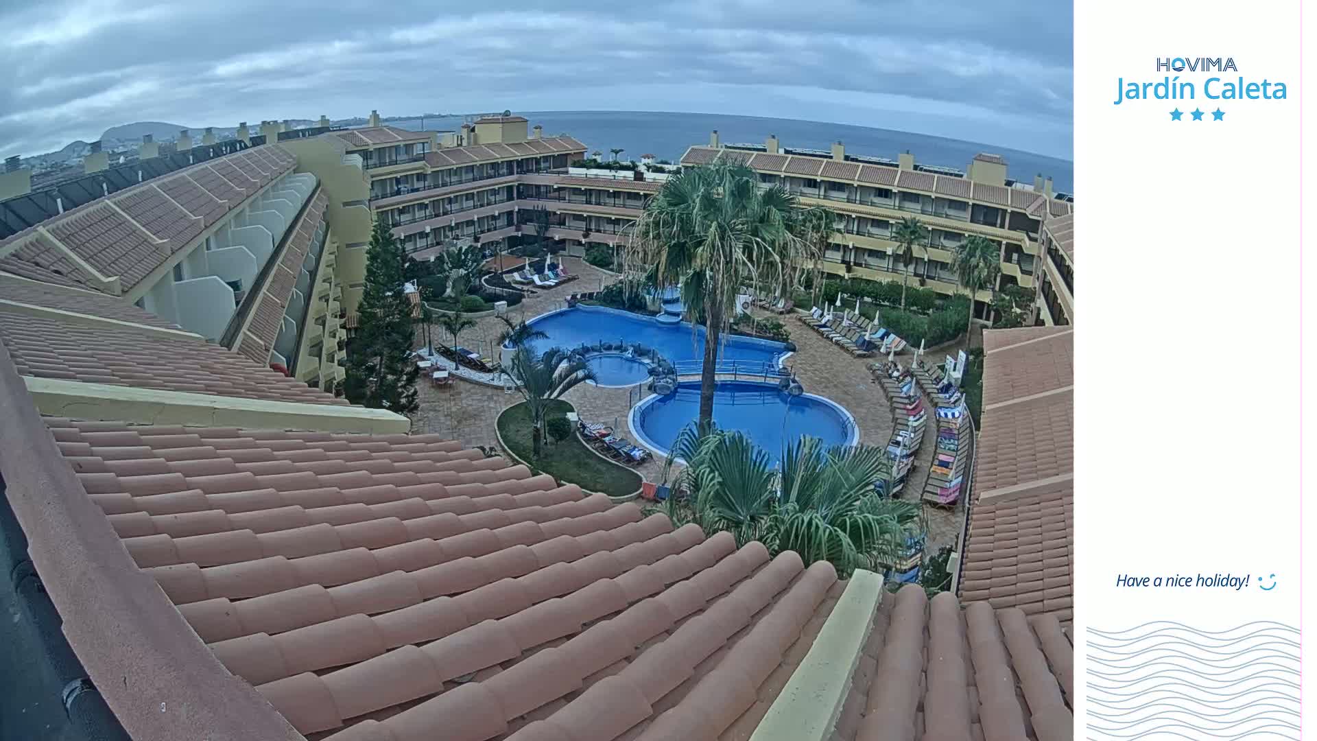 An aerial view of a resort complex with a swimming pool, palm trees, and tiled rooftops, under a cloudy sky.
