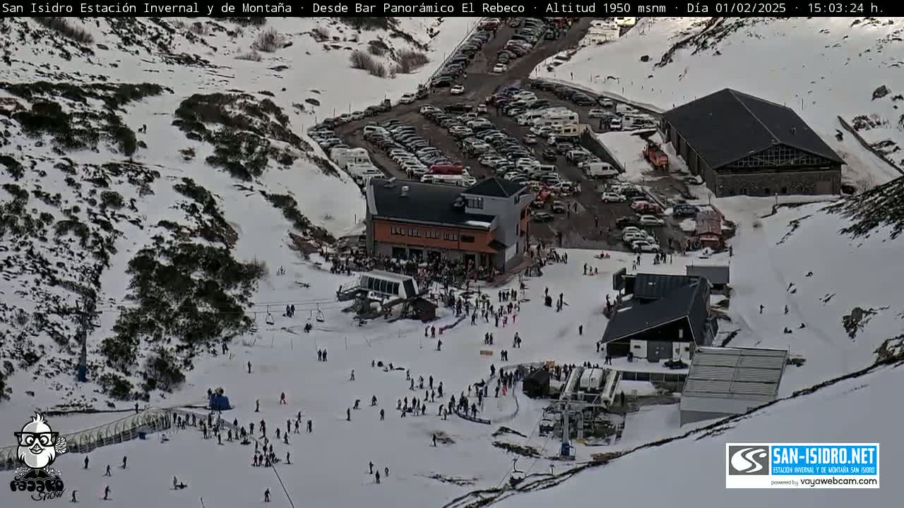 A snowy mountain ski resort with several buildings, a large parking lot full of cars, and many people skiing and snowboarding on the slopes.