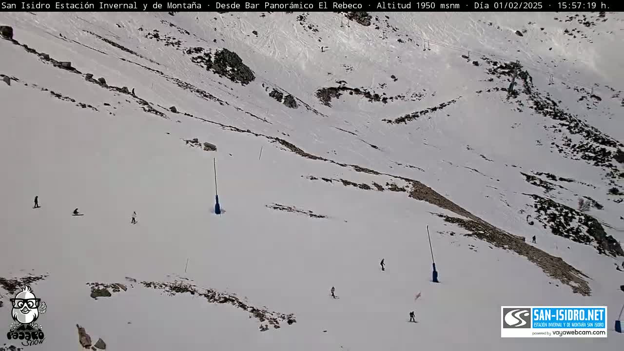 A snowy ski slope with several skiers and snowboarders visible, under a bright, sunny sky.