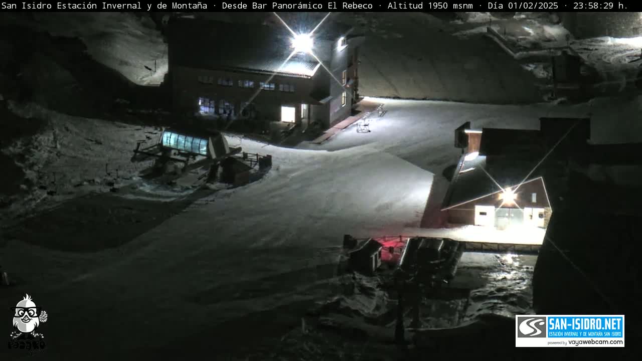 A snow-covered ski resort at night, illuminated by artificial lights, showing buildings and ski lifts.
