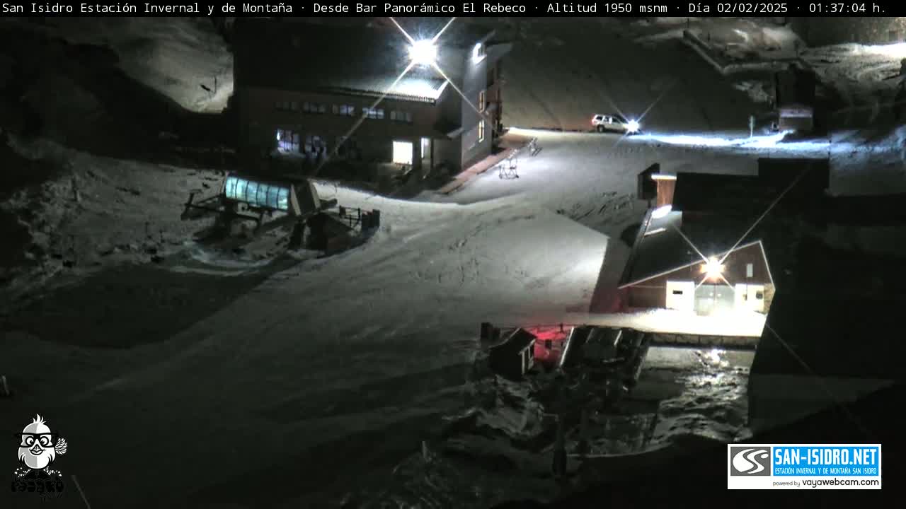 A snow-covered ski resort at night, illuminated by artificial lights, shows several buildings and a ski lift.