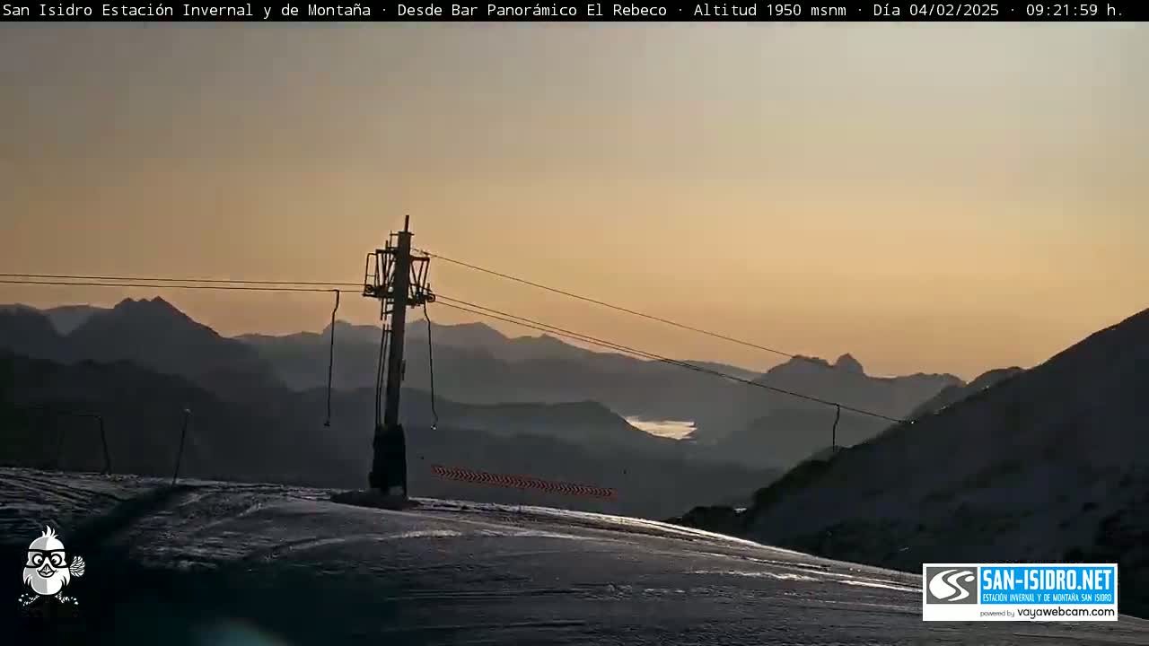 A ski lift stands atop a snow-covered mountain slope at sunset, overlooking a valley and distant, silhouetted mountain ranges.