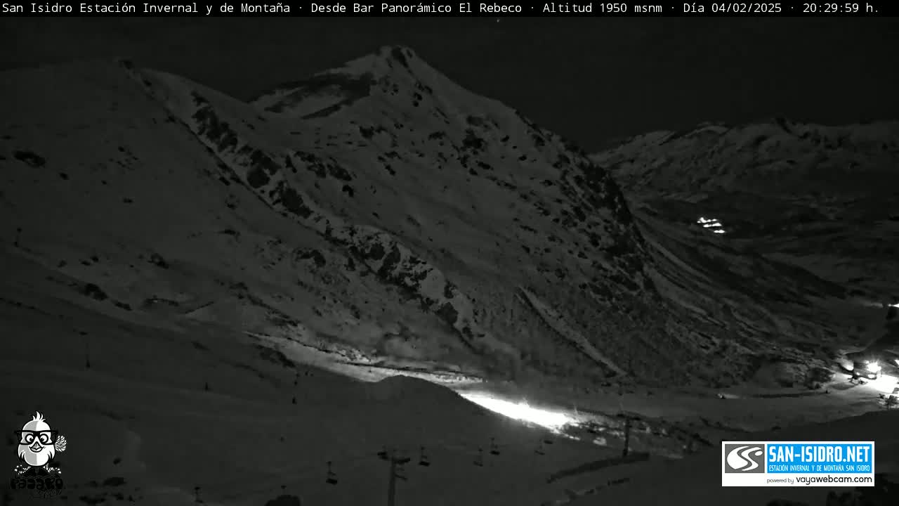 A nighttime, low-light image shows a snow-covered mountain slope with some lights visible in the valley below.