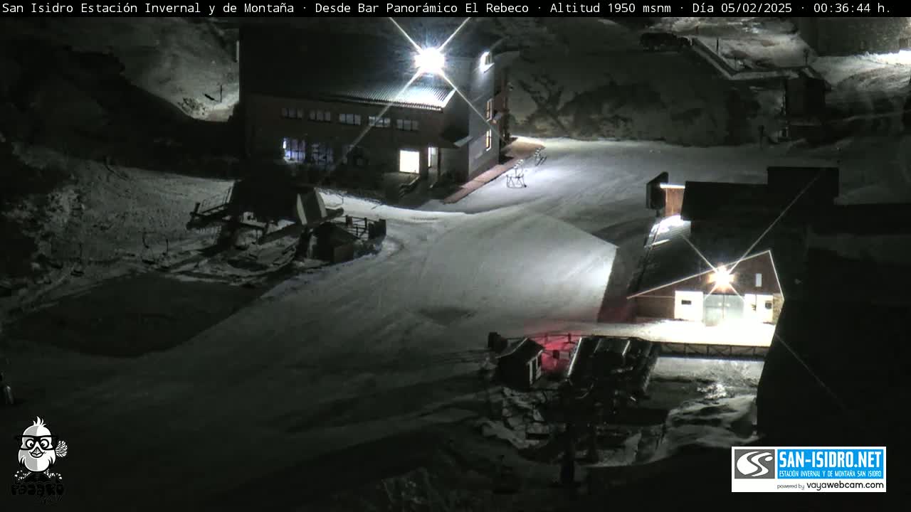 A nighttime, snow-covered ski resort scene shows two buildings illuminated, with snow-grooming equipment visible near one.