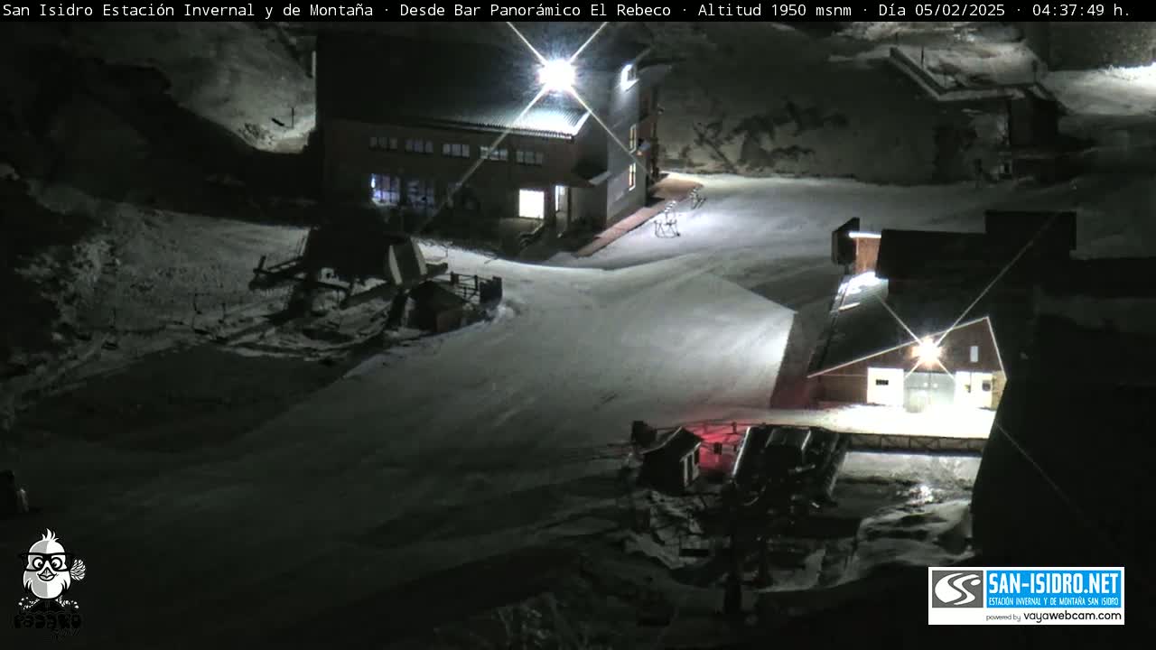 A nighttime, snow-covered mountain resort scene shows several buildings illuminated under artificial light.