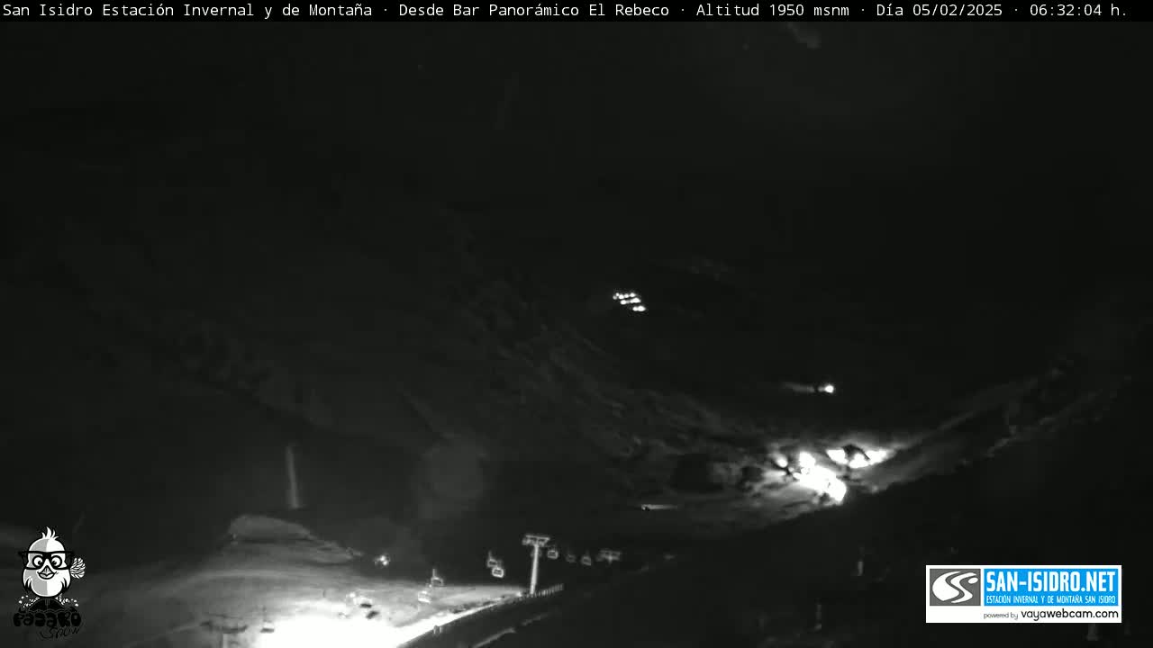 A nighttime, low-light view of a snow-covered mountain slope with illuminated ski lifts and buildings.