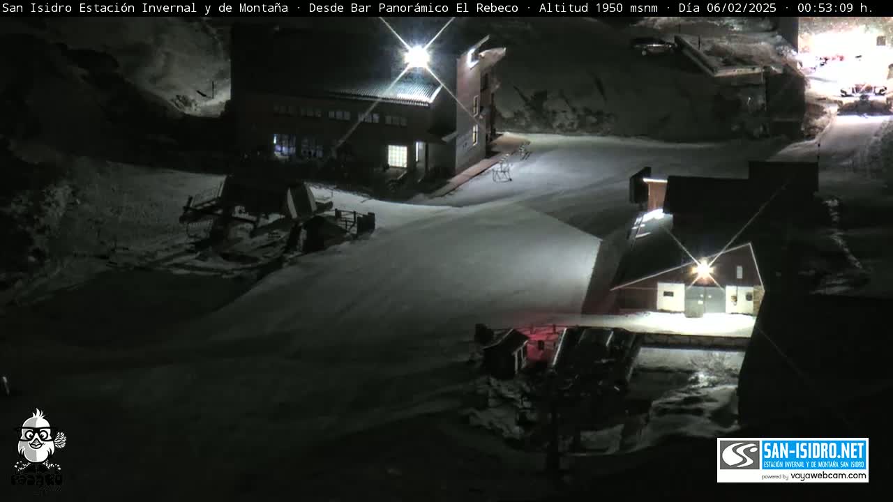 A nighttime snow scene at a ski resort shows several buildings and snow-grooming equipment under bright lights.