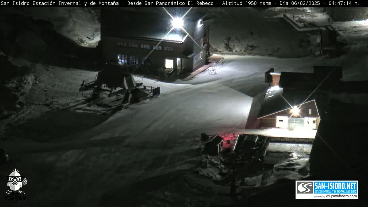 A snow-covered nighttime scene shows several buildings, snow-grooming equipment, and a mostly empty roadway.