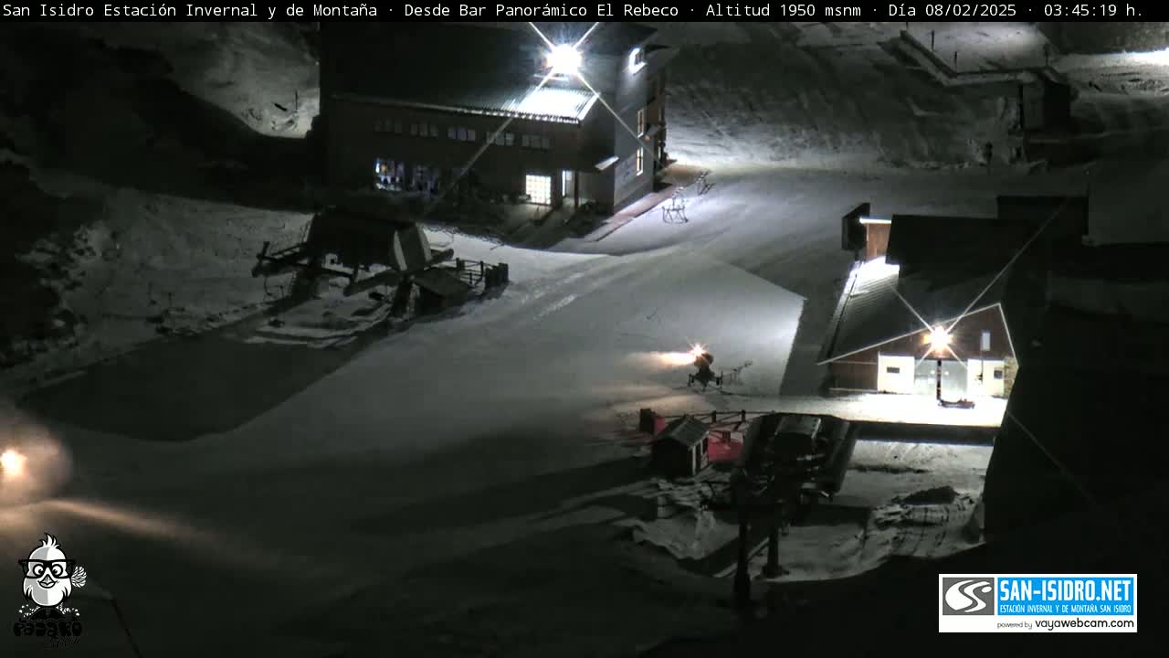 A snow-covered ski resort at night, illuminated by artificial lights, with snow groomers and ski lifts visible.
