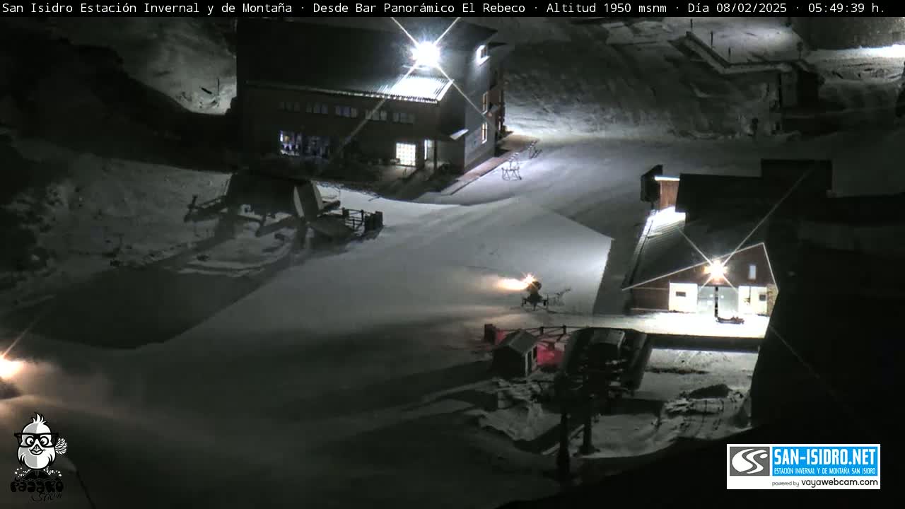 A snow-covered nighttime scene shows a snowmaking machine operating near two buildings, illuminated by lights.