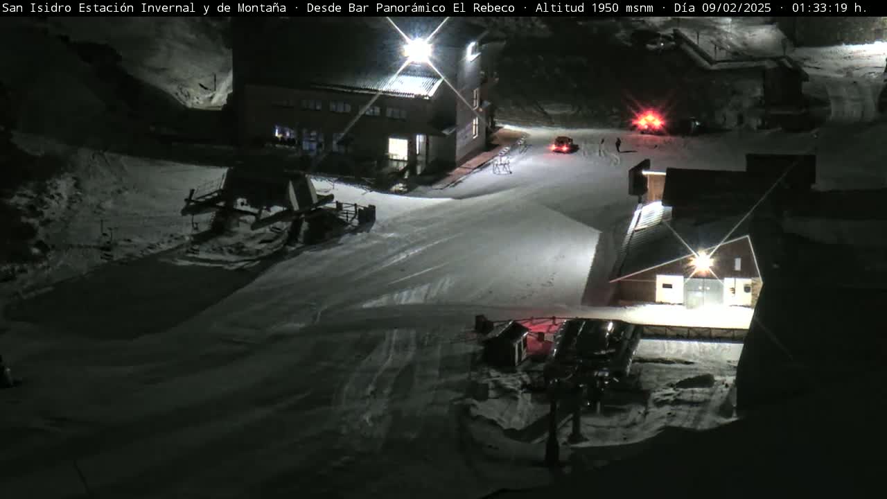 A snow-covered ski resort at night, illuminated by artificial lights, shows buildings, ski lifts, and snowcats.