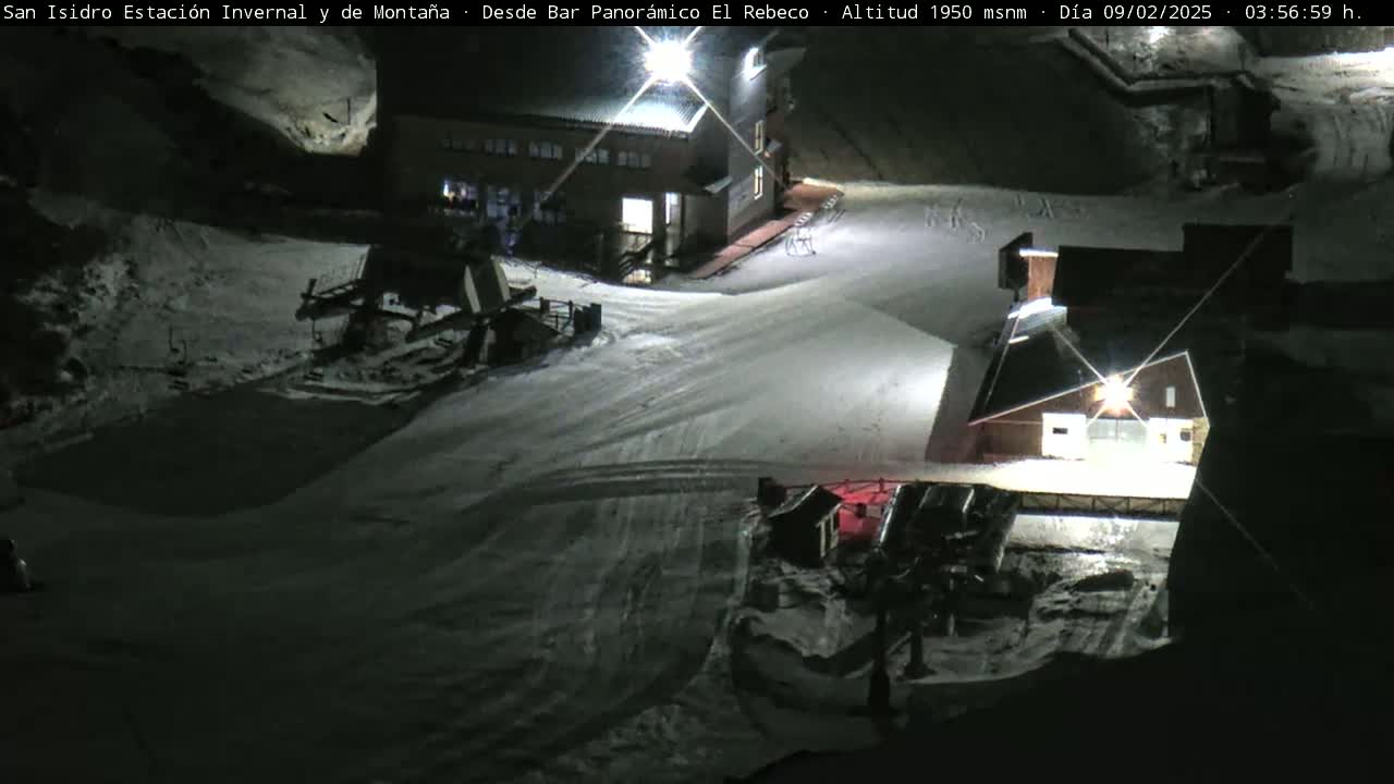 A snow-covered ski resort at night, illuminated by artificial lights, showing several buildings and snow grooming equipment.