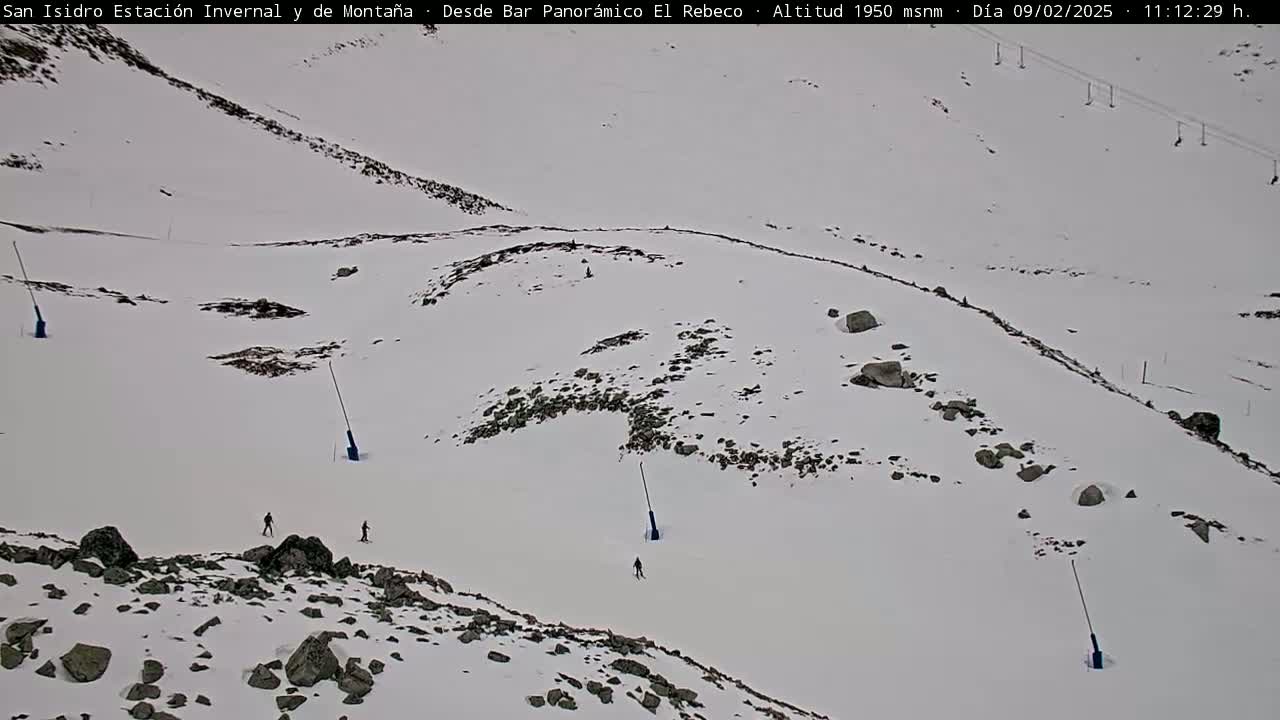 A snowy, rocky mountain slope with a few skiers and ski lift towers visible under an overcast sky.