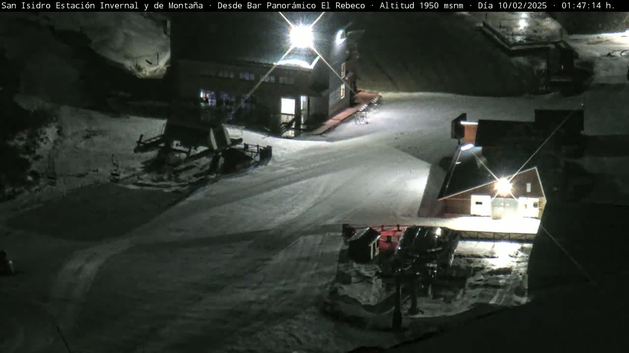 A snow-covered ski resort at night, illuminated by artificial lights, with several buildings and snow-grooming machinery visible.