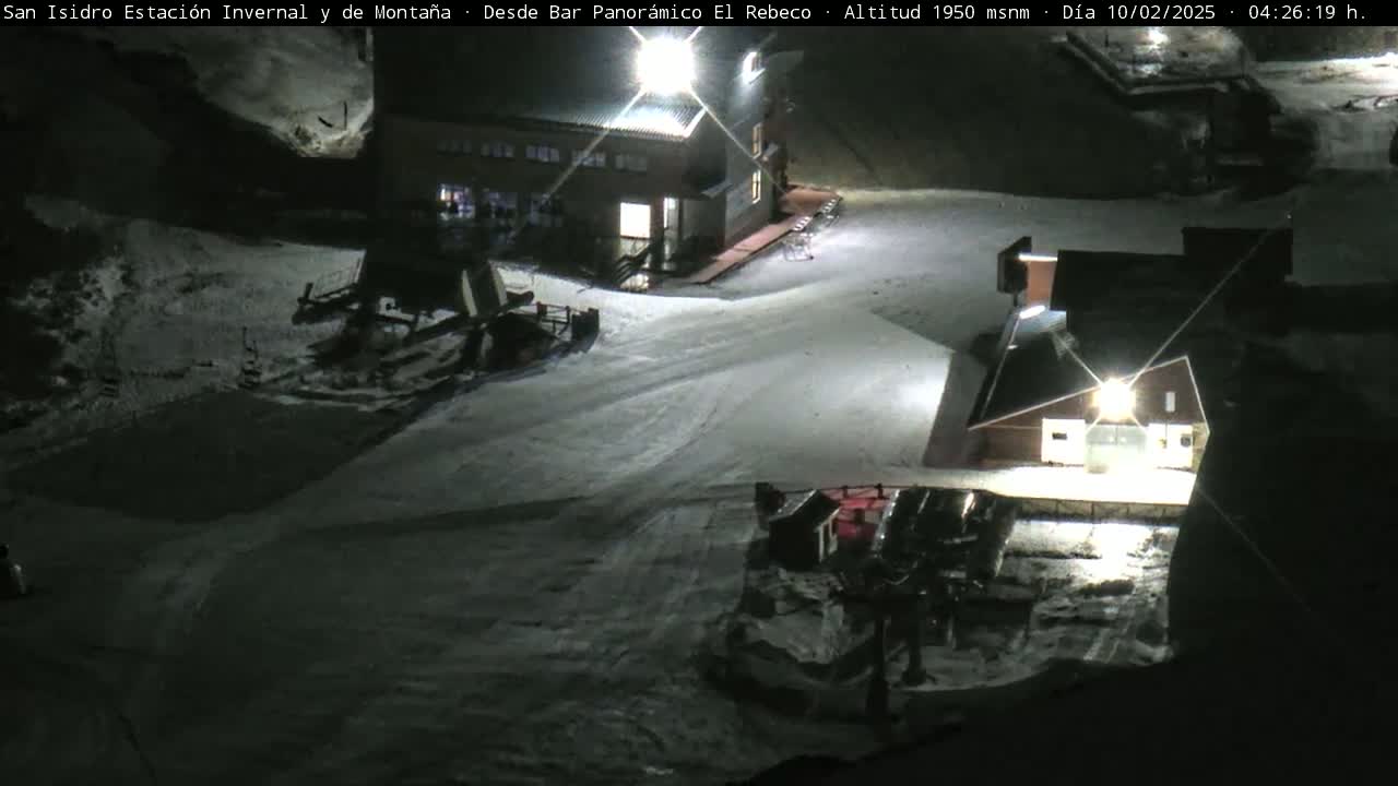 A snow-covered ski resort at night, illuminated by artificial lights, showing several buildings and snow grooming equipment.