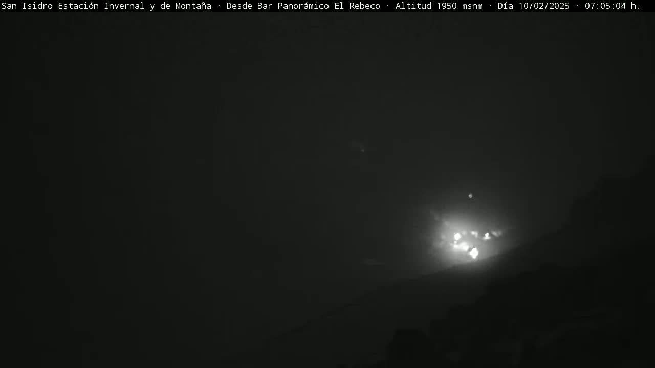 A nighttime, dimly lit outdoor scene shows a bright light source in the distance, possibly from a building or vehicle, with a dark, hazy or foggy atmosphere.