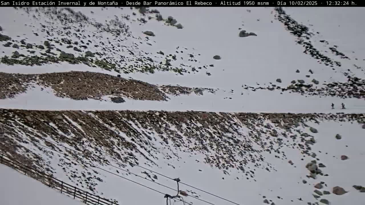 A snowy mountain slope with dark rocks and patches of exposed ground, a ski lift, and two figures skiing in the distance.