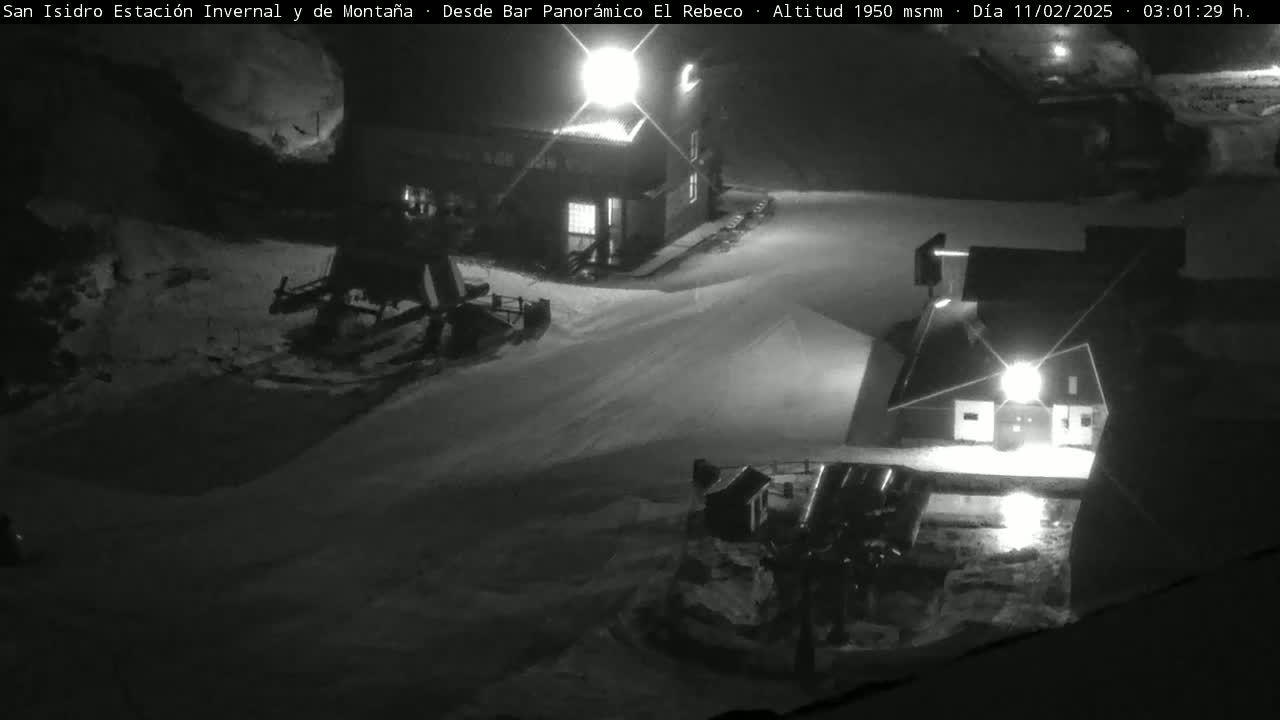 A nighttime, snow-covered mountain scene shows two illuminated buildings and some equipment, possibly ski lifts.