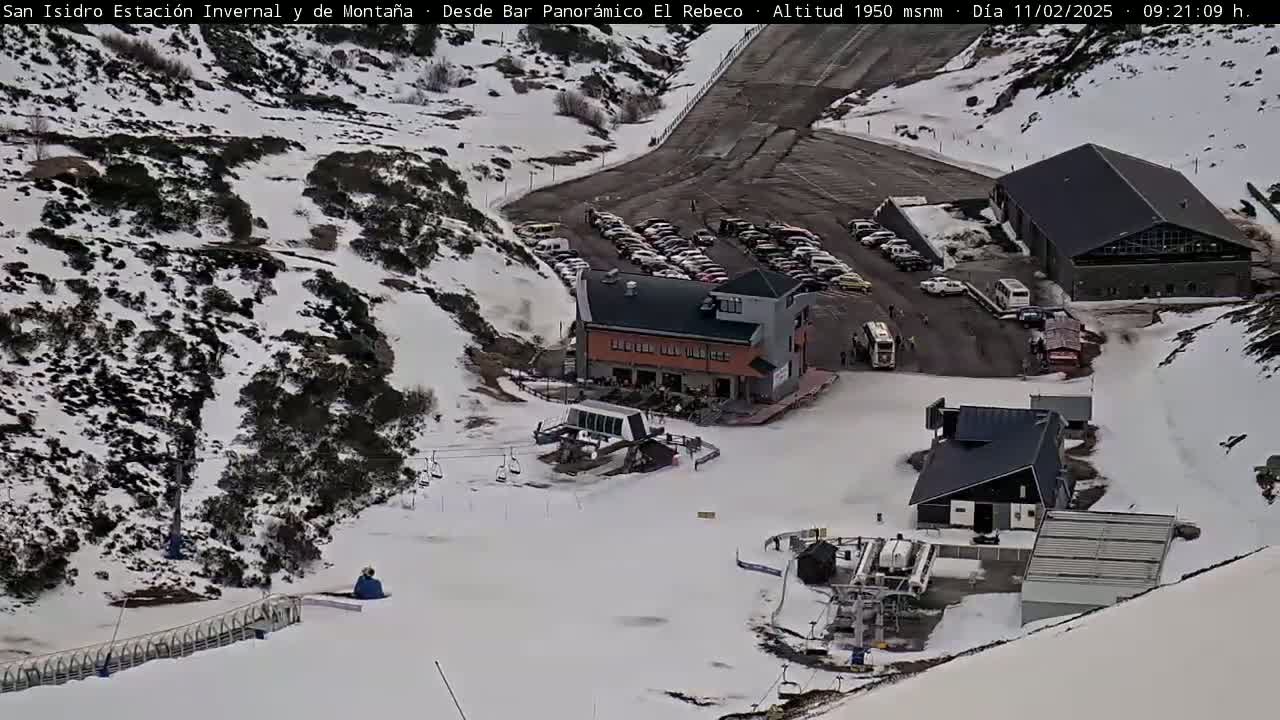 A snow-covered mountain slope with ski lifts, buildings, and a parking lot full of cars.