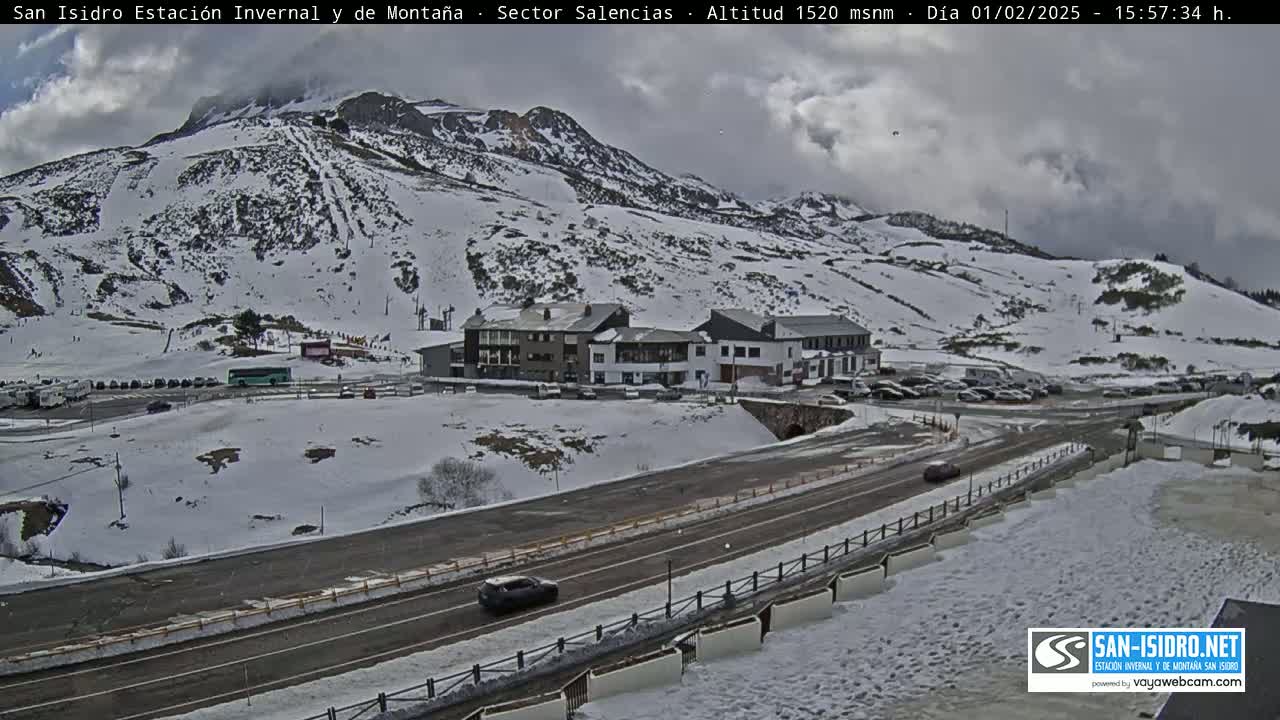 A snow-covered mountain slope with a resort building and a road below, under a cloudy sky.