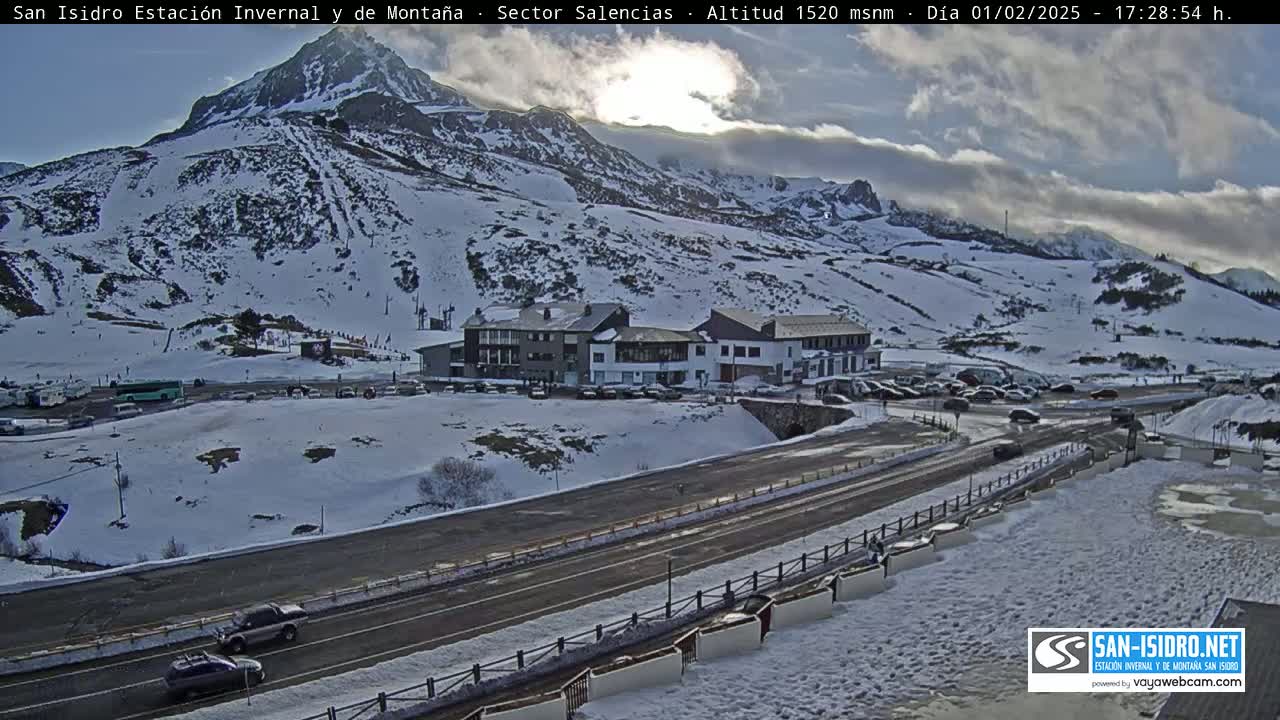 A snow-covered mountainside with a road and buildings, under a partly cloudy sky.