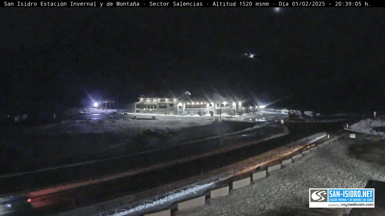 A nighttime, snow-covered landscape shows a large building illuminated near a highway.