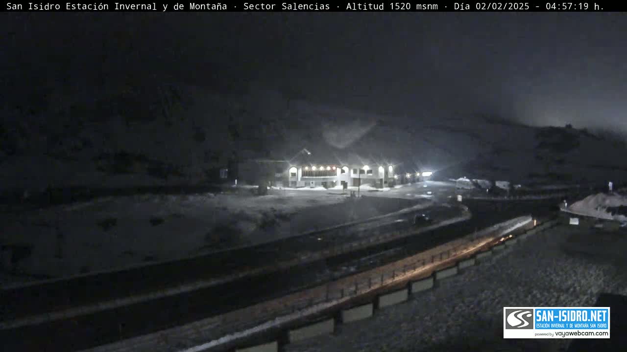 A snow-covered mountain landscape at night, featuring a building illuminated in the foreground and a roadway, under dark, possibly cloudy, conditions.