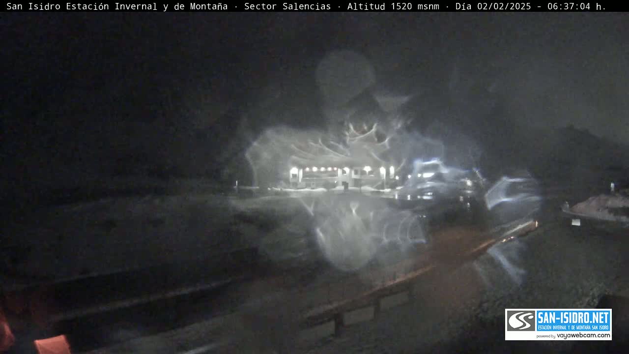 A nighttime, snowy scene shows a building and several vehicles, with blurry, streaking lights suggesting blowing snow or rain.