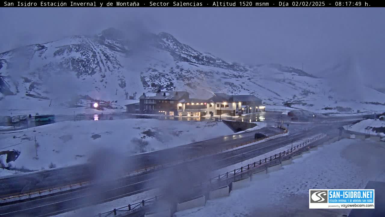 A snow-covered mountain landscape at dusk features a roadway leading to a building complex, with light snow falling.