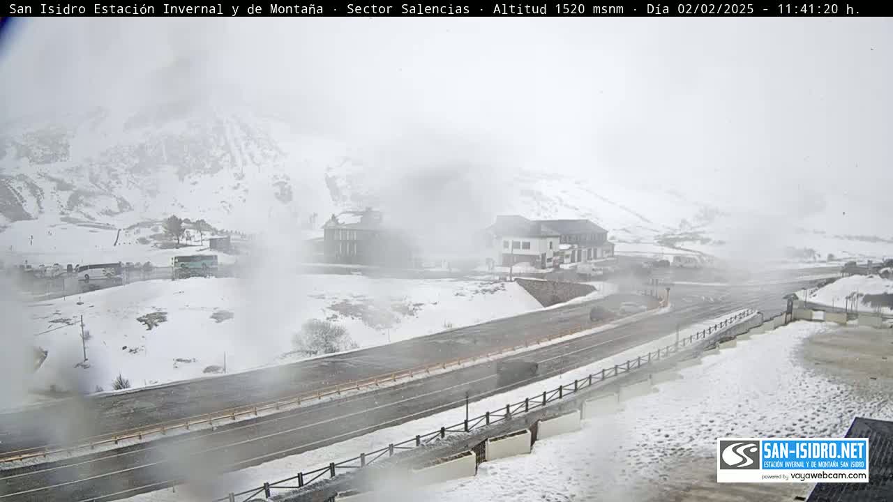A snow-covered mountain road winds past buildings in a hazy, snowy landscape.