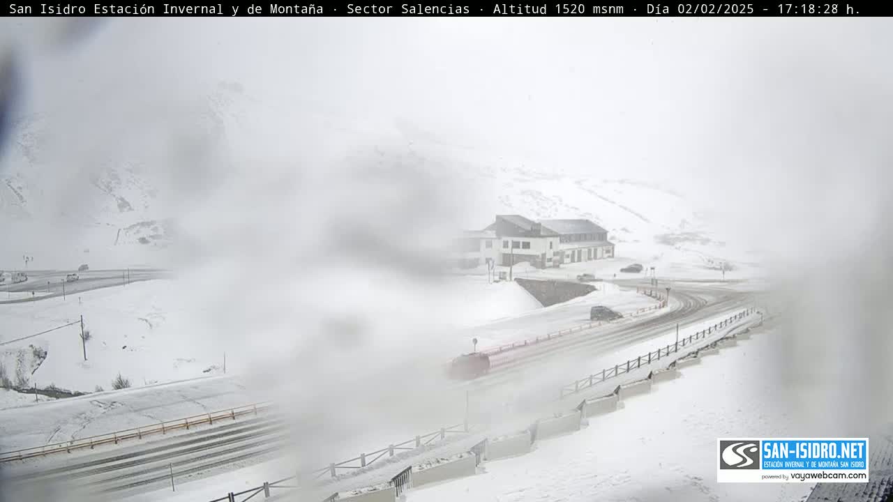 A snow-covered mountain road winds past a building, with blowing snow obscuring the view.