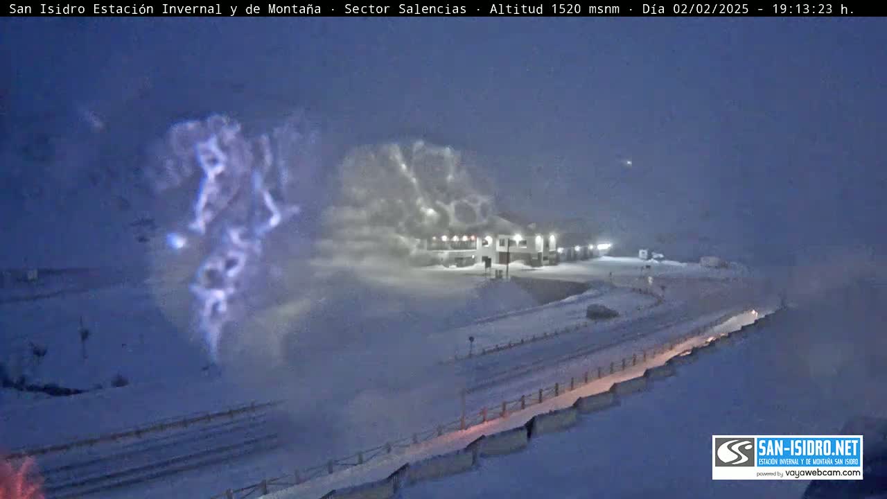 A snow-covered landscape at night, with a building illuminated and a large plume of snow blowing in the foreground under a twilight sky.