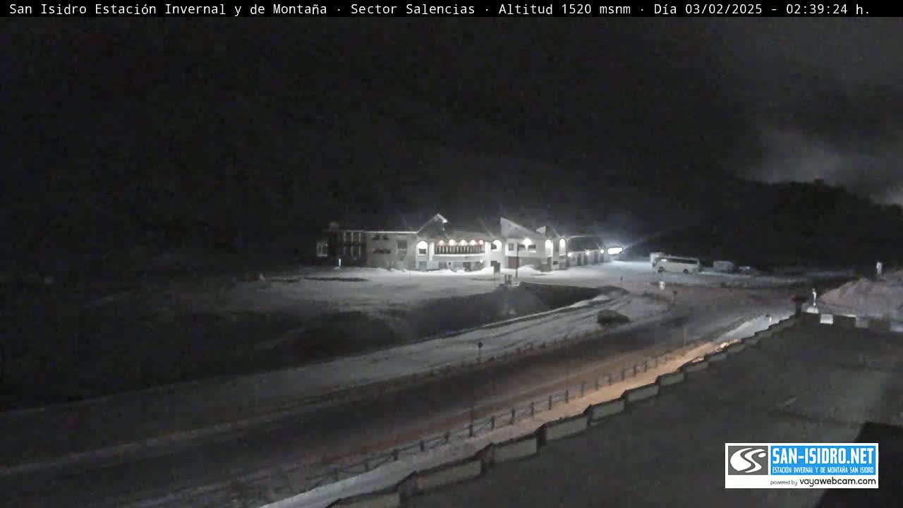 A snow-covered parking lot and roadway in front of a lit building at night.