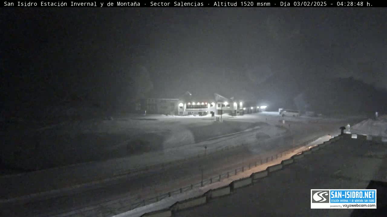A snow-covered landscape at night, showing a building and a road under heavy snowfall.