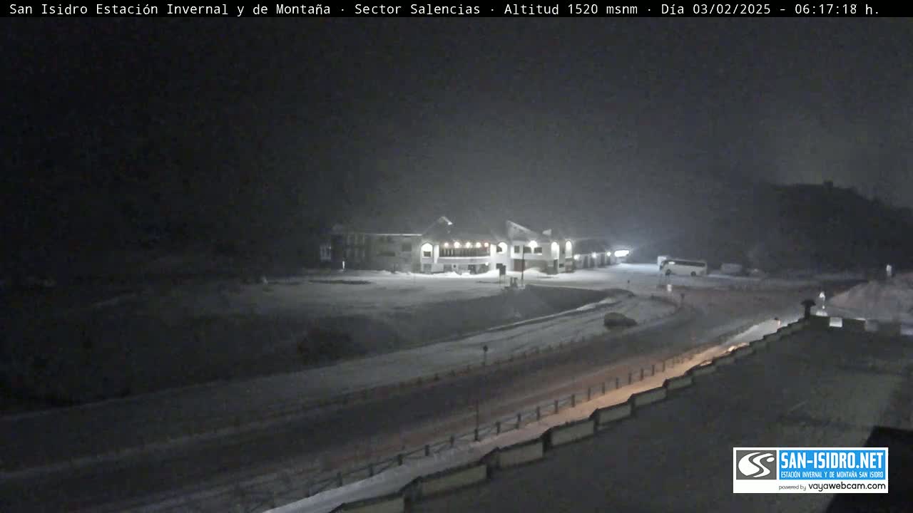 A snow-covered landscape at night shows a building illuminated, with a road and a few vehicles visible.