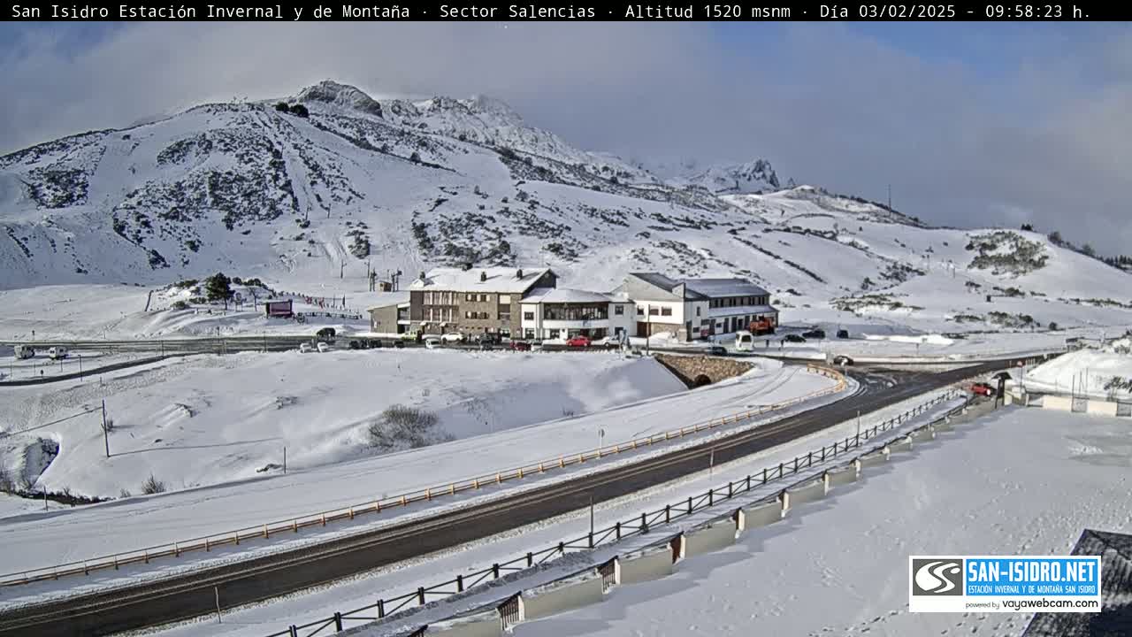 A snow-covered mountain landscape with a road, buildings, and a few vehicles, under a mostly clear sky.