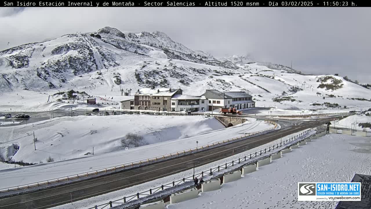 A snow-covered mountain slope with ski runs, a road, and several buildings is visible under a cloudy sky.