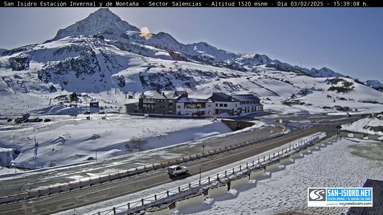 A sunny, snow-covered mountain landscape with a road, a building complex, and a few cars.