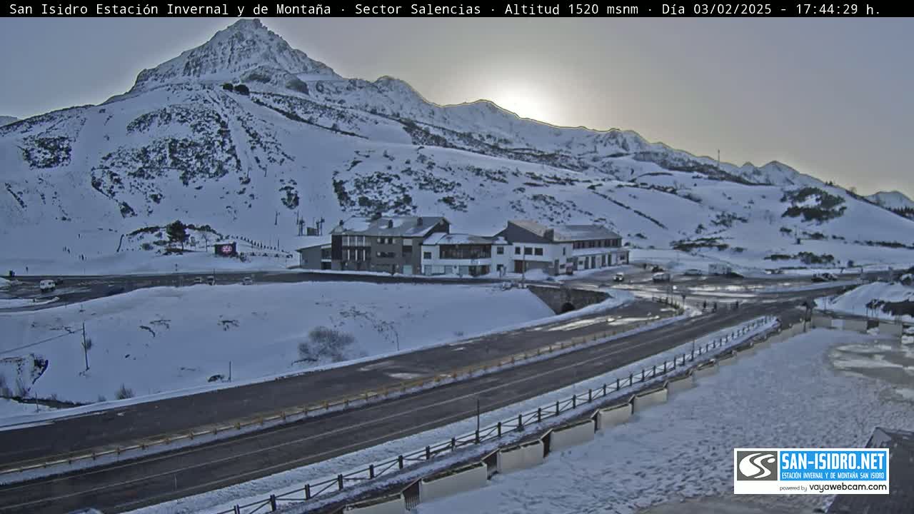 A snow-covered mountainside with a few buildings and a road, under a partly sunny sky.