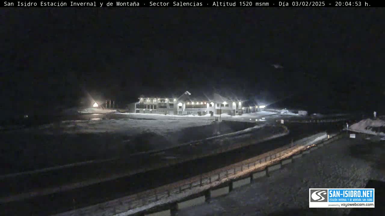 A snow-covered landscape at night shows a lit building near a road.
