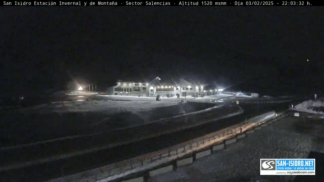 A snow-covered nighttime scene shows a large building illuminated, with a road and fencing visible in the foreground.