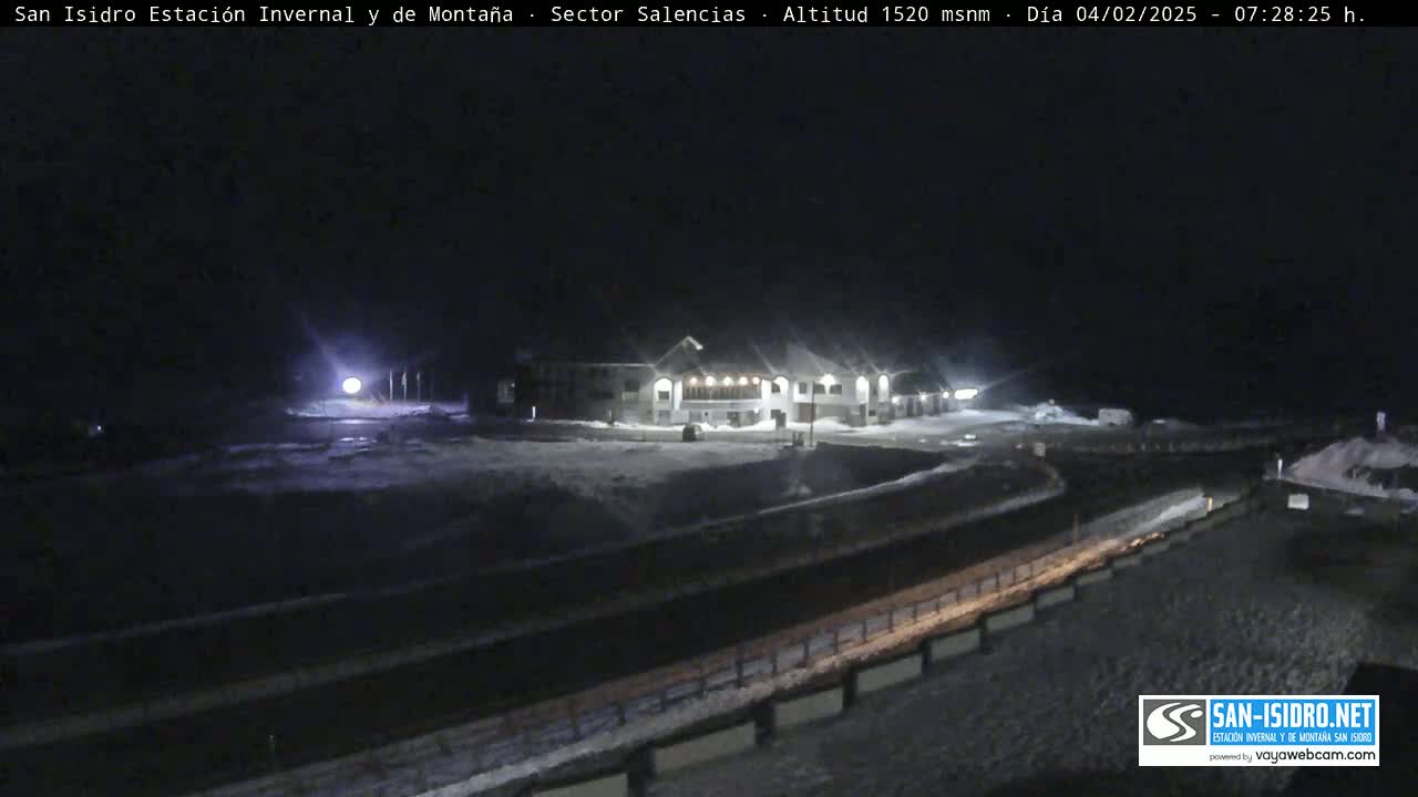 A snow-covered nighttime scene shows a building complex illuminated by lights, with a road and snow-covered ground in the foreground.