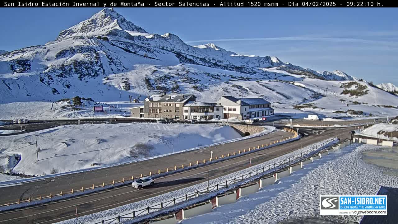 A snow-covered mountain landscape with a road winding past a building complex, under a clear blue sky.