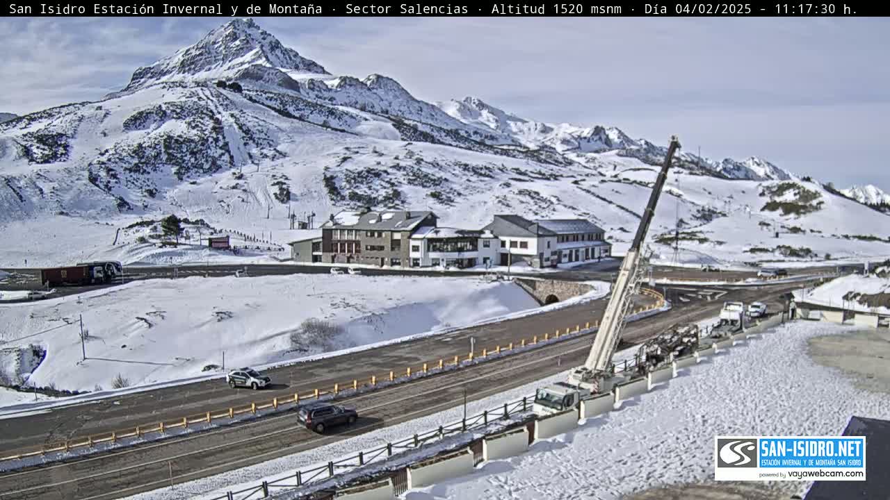 A snow-covered mountain landscape with a road, buildings, and several vehicles, under a mostly clear sky.
