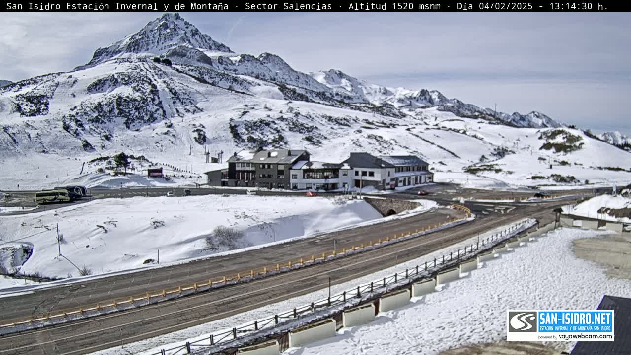 A snow-covered mountainside with a road and buildings below, under a mostly clear sky.